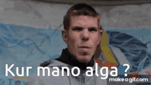 a man is giving the middle finger in front of a wall that says kur mano alga .