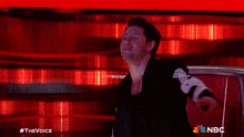 a man in a black jacket is standing in front of a red wall with nbc written on the bottom right