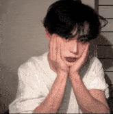 a young man with his hands on his face is wearing a white shirt and looking at the camera .