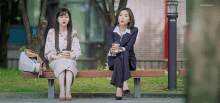 two women are sitting on a park bench drinking coffee