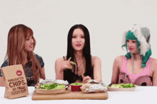 three women are sitting at a table eating chips from a bag that says handcrafted chips