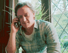 a man in a plaid shirt is talking on a cell phone in front of a window