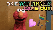 elmo from sesame street says " okie you finally came out " while holding a heart and rainbow flags