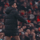 a man in a black jacket stands in front of a crowd with the letters x on the bottom left