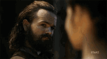a man with a beard and long hair is standing next to a woman and looking at her .