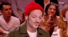 a man wearing a red beanie and a green jacket is sitting in a crowd of people .