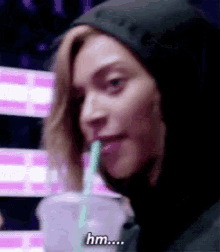 a woman in a hoodie is drinking through a straw and says hm .