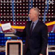 a man in a suit and tie holds a piece of paper in front of a sign that says family feud canada