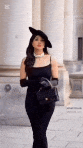 a woman in a black dress , hat , gloves and a purse is walking down the street .