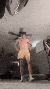 a shirtless man in yellow shorts is standing in a living room with a ceiling fan in the background