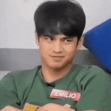a young man wearing a green shirt with a name tag that says emilio is sitting with his arms crossed .