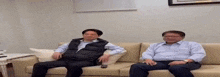 two men are sitting on a couch in a living room with a remote control in their hand .