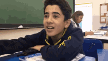 a boy sits at a desk in a classroom wearing a sweatshirt with the letter g on the sleeve