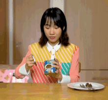 a woman sitting at a table with a plate and spoon holding a can of beans