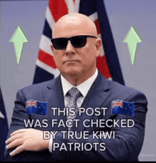 a bald man wearing sunglasses and a suit is standing in front of a british flag and a kiwi flag .