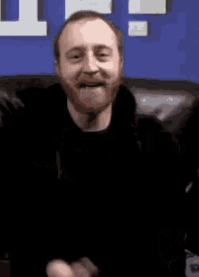 a man with a beard is sitting on a couch making a funny face and giving the middle finger .