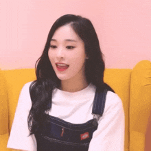 a girl wearing overalls and a white shirt is sitting on a yellow couch .