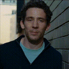 a man standing in front of a brick wall wearing a black sweater and a blue shirt