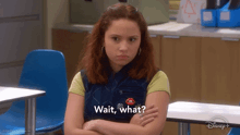 a girl is sitting at a desk with her arms crossed and says " wait what "