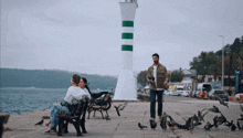 a man standing next to a woman sitting on a bench in front of a green and white lighthouse