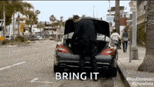 a man is putting something in the trunk of a car and the words bring it are visible