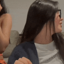 two women are sitting on a couch talking to each other . one of the women is wearing glasses .