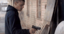 a man in a black hoodie is holding a gun in front of a brick building .