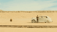 two men standing next to a white car in the middle of the desert