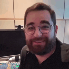 a man with glasses and a beard smiles in front of a computer monitor