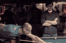 a man in a blue shirt is playing pool while another man watches
