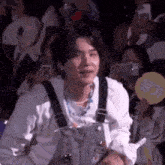 a man in overalls is standing in front of a crowd of people holding a fan and a cell phone .