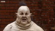 a statue of a monster with a mohawk is standing in front of a brick wall and making a funny face .