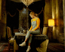 a woman in overalls sits on a table in a room with a lamp
