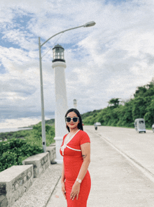 a woman in a red dress and sunglasses stands in front of a lighthouse