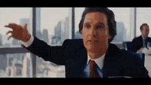 a man in a suit and tie is sitting at a table with his arms outstretched in front of a window .