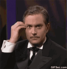 a man in a tuxedo and tie is making a funny face with his hand in the air .