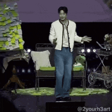 a man in a white jacket and blue jeans is standing on a stage with his arms outstretched .