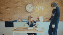 a woman sits at a desk in front of a clock with roman numerals and a man standing next to her