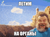 a man with a beard and a blue shirt is making a funny face while standing in front of a mountain .