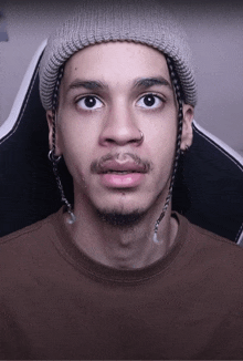 a young man wearing a beanie and a brown shirt looks at the camera with a surprised look on his face
