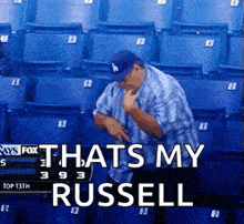 a man in a baseball cap is sitting in a stadium with the words that 's my russell on the bottom .