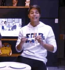 a young man wearing a white star wars hoodie is sitting at a table with his hands in the air .