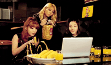 three girls are looking at a laptop and one of them has a bag of corn on the cob in her hand