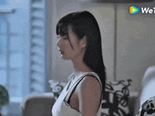 a woman in a white tank top is standing in front of a window with a play button in the corner .