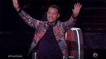 a man in a colorful jacket is sitting in a chair with his arms in the air and a nbc logo behind him