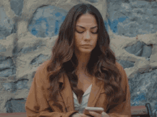 a woman with long hair is sitting on a bench in front of a stone wall with the letter g on it
