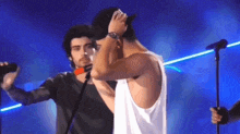two men are singing into microphones on a stage . one of the men is wearing a hat .
