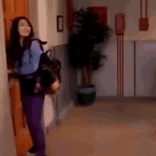 a woman in purple pants is standing in a hallway holding a backpack .