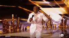 a man in a white suit is holding a microphone and dancing on a stage with a nbc logo behind him