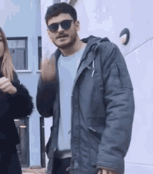 a man wearing sunglasses and a jacket is standing next to a woman and waving .